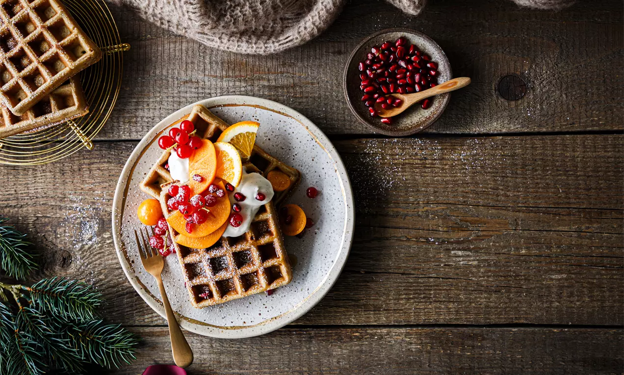 Picture - Square golden-brown waffles topped with cream, orange slices, red pomegranate seeds and kumquats on a speckled plate with a fir sprig