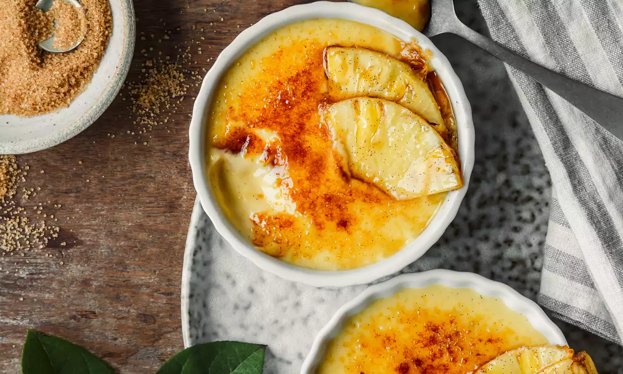 Picture - Coconut creme brulee in white ramekins with golden caramelised tops, garnished with pineapple slices on a speckled plate with green leaves