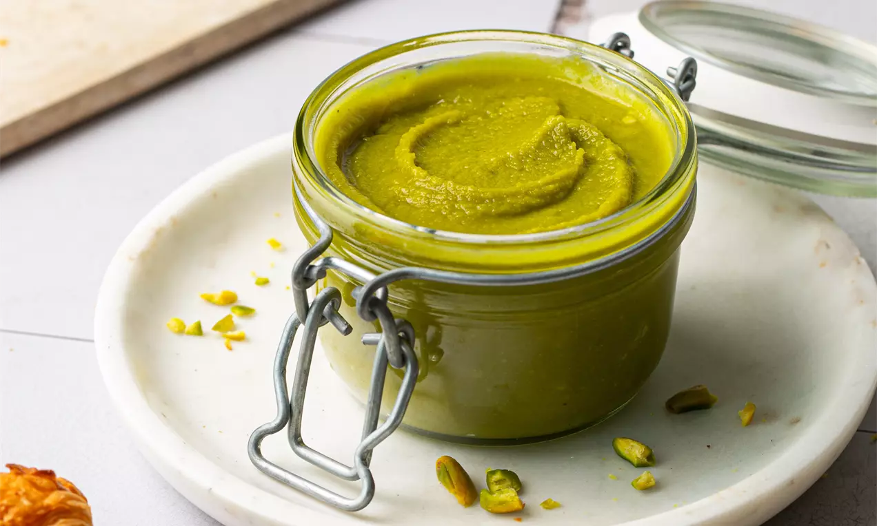 Picture - A glass jar of smooth, vibrant green pistachio cream with swirls, placed on a white plate, scattered pistachios and golden croissants nearby