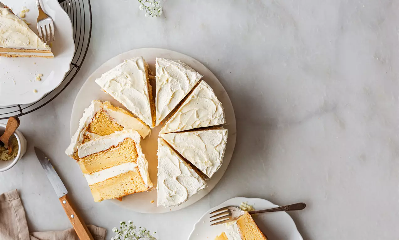 Picture - Golden vanilla cake sliced into triangular pieces, layered with pale cream frosting, arranged on a round plate on a marble surface