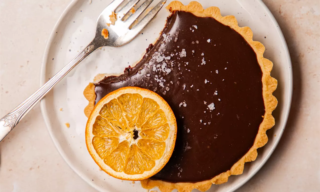 Picture - Golden tart shell filled with glossy dark chocolate, garnished with a thin orange slice and a sprinkle of salt on a white plate