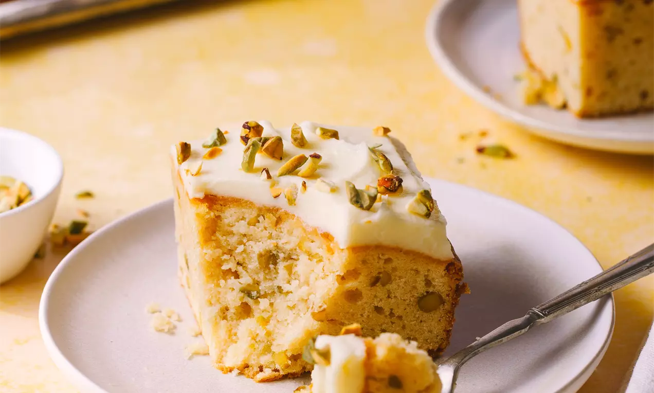 Picture - A square slice of pale golden lemon blondie topped with creamy glaze and chopped pistachios, on a white plate with a fork