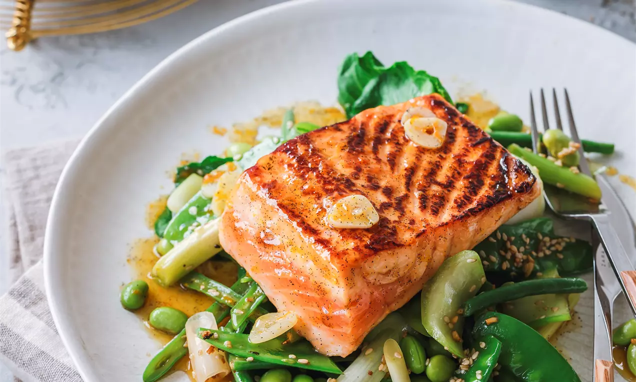 Picture - Grilled salmon fillet with a golden crust atop vibrant green vegetables, drizzled with a light sauce on a white plate