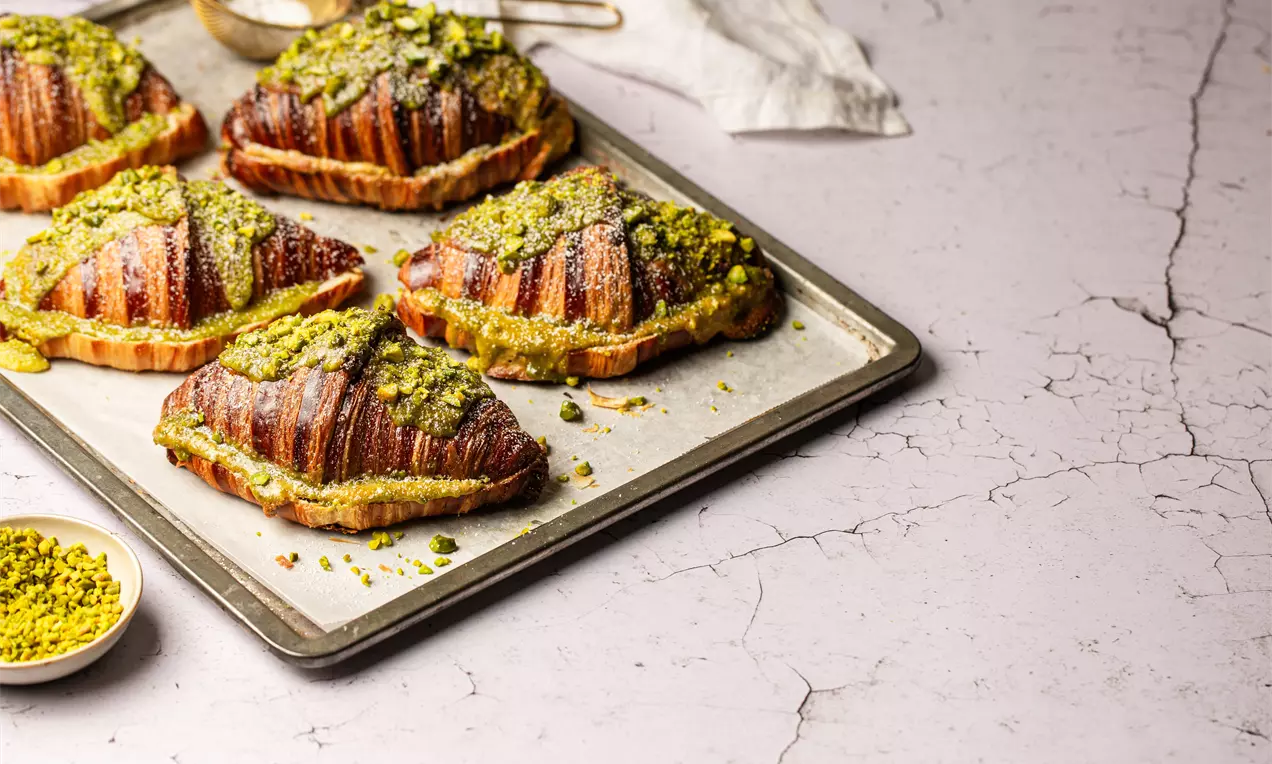 Picture - Golden brown croissants with crisp layers, topped with crushed green pistachios and powdered sugar, arranged on a metal tray