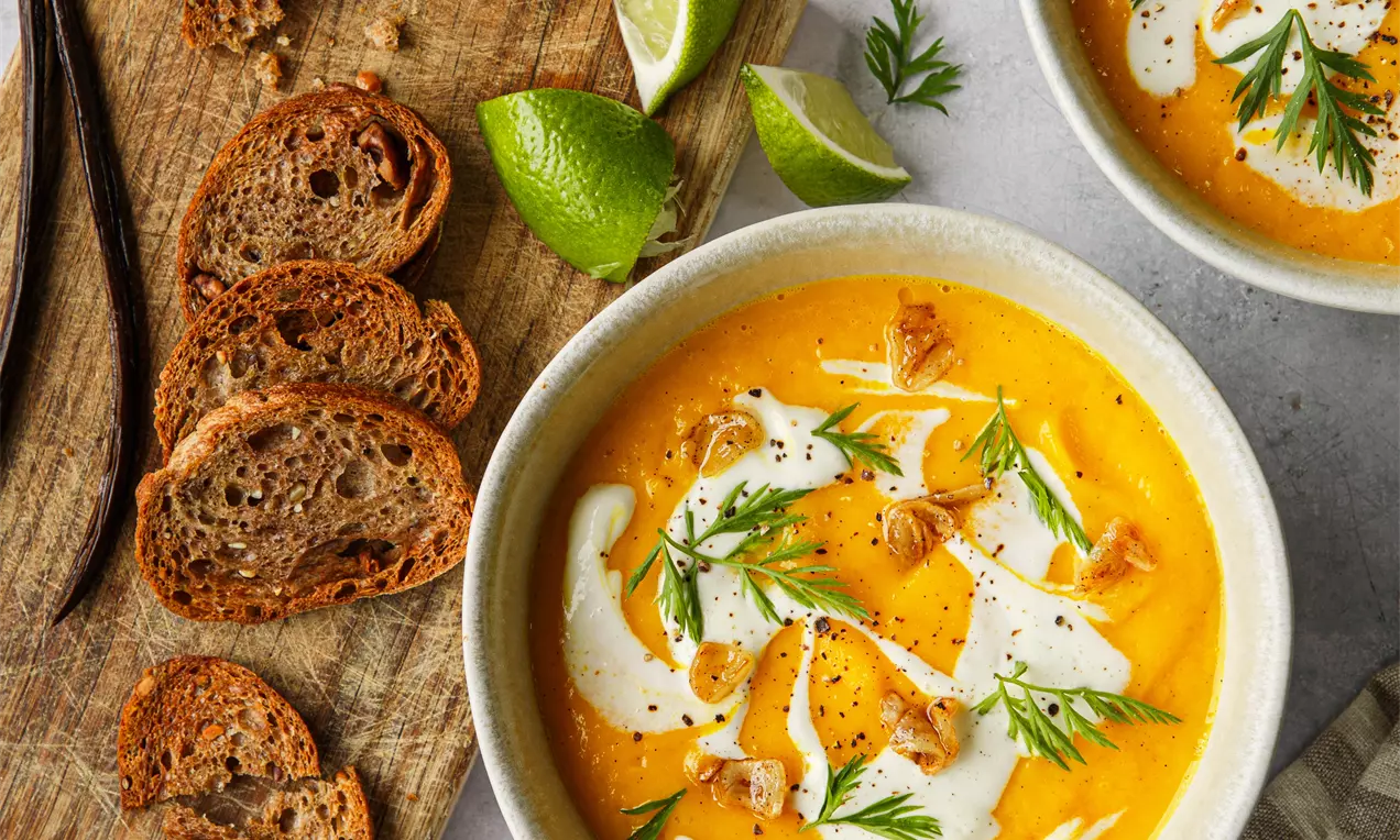 Picture - Vibrant orange carrot soup in ceramic bowls topped with white cream swirls, fresh herbs, and croutons on a textured surface